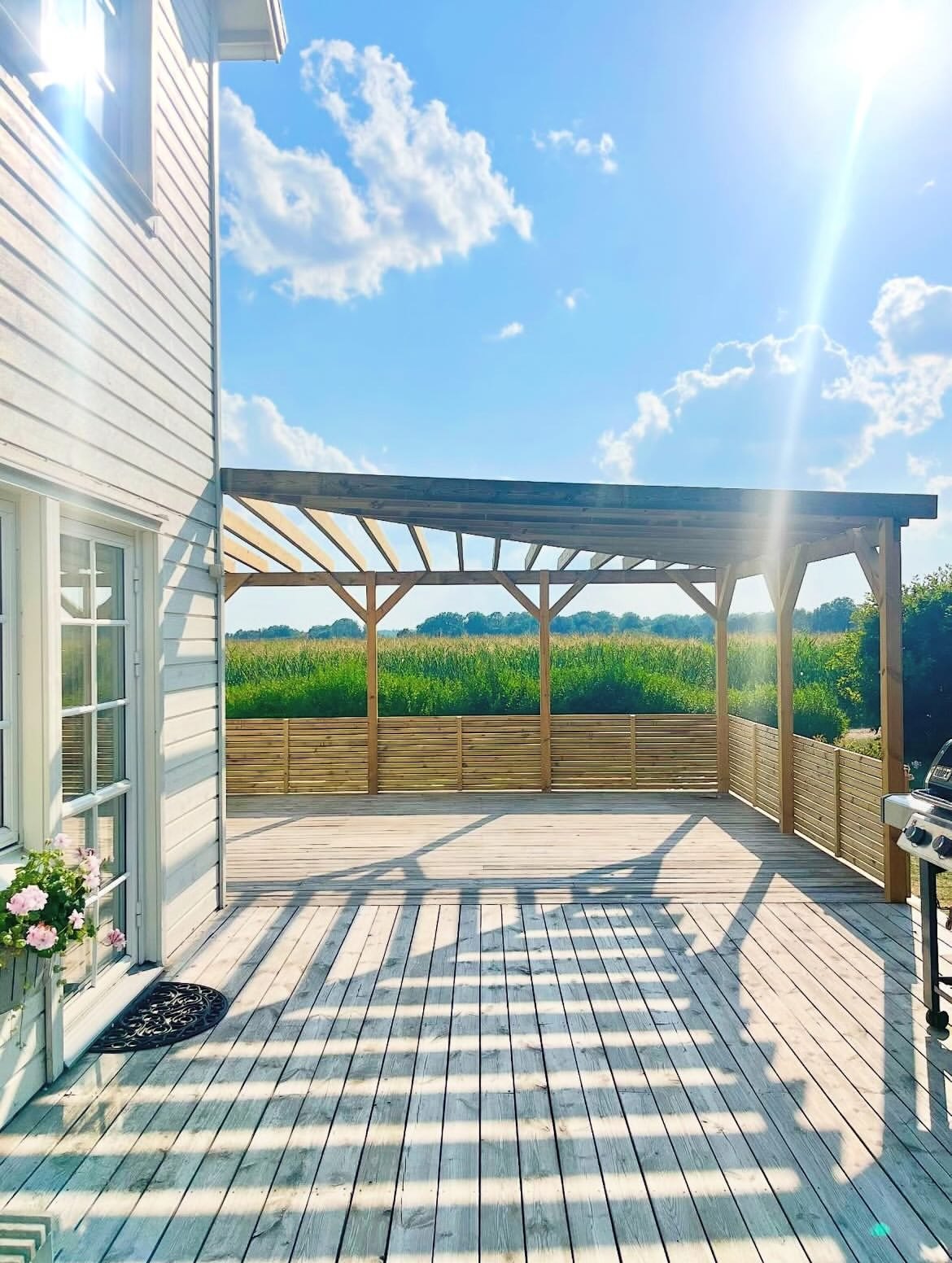 Pergola i obehandlat trä över en altan med insynsskydd och utsikt över fält