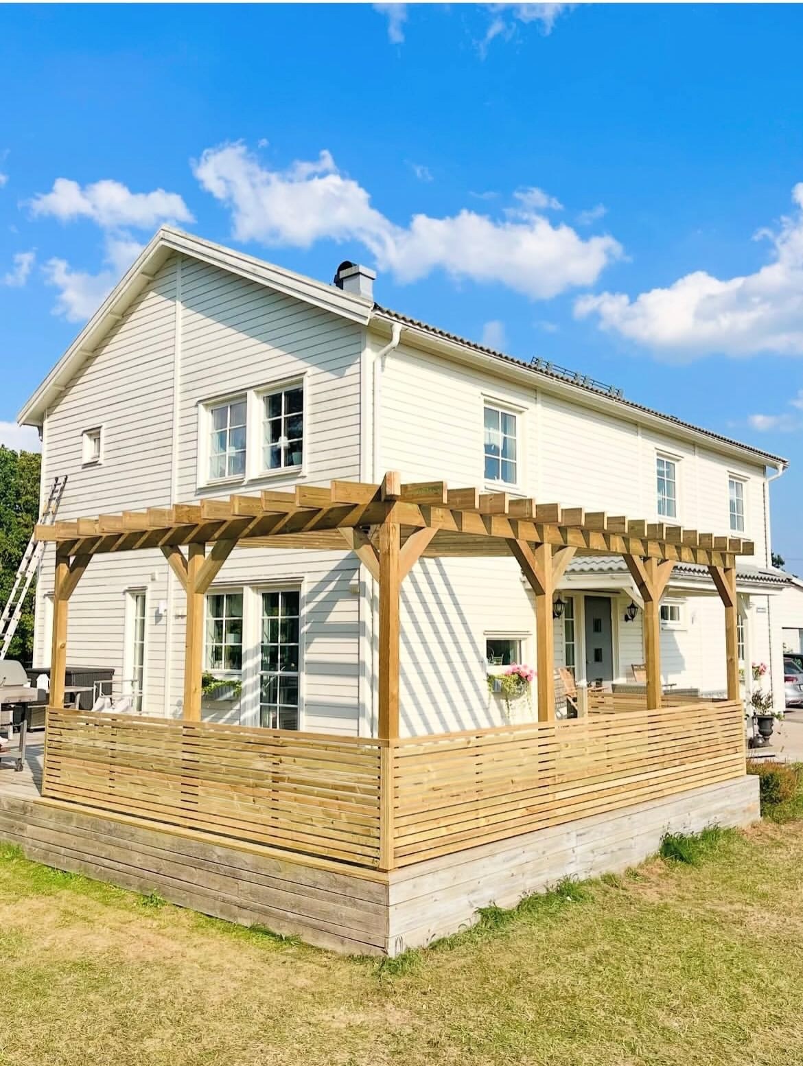 Pergolan sedd utifrån mot det vita huset med altan och insynsskydd i obehandlat trä