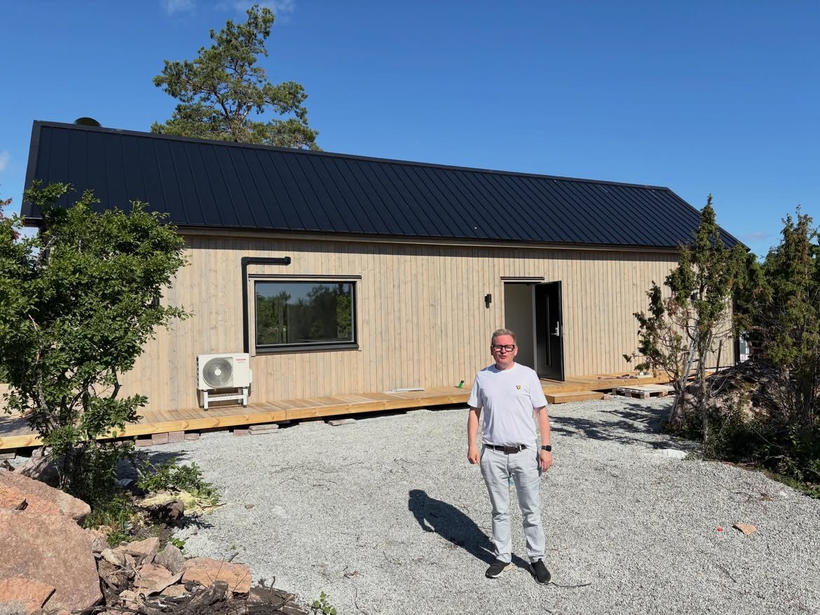 Nybyggt fritidshus med ljus träpanel, svart falsat plåttak och trädäck runt entrén